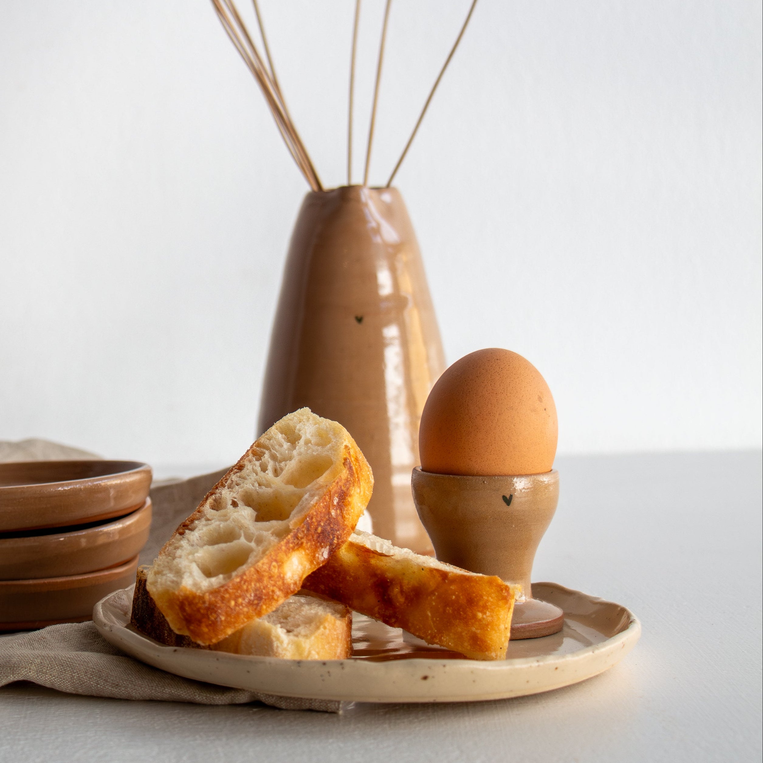 Ceramic vase with dried plants, plates with bread and an egg on a light surface.