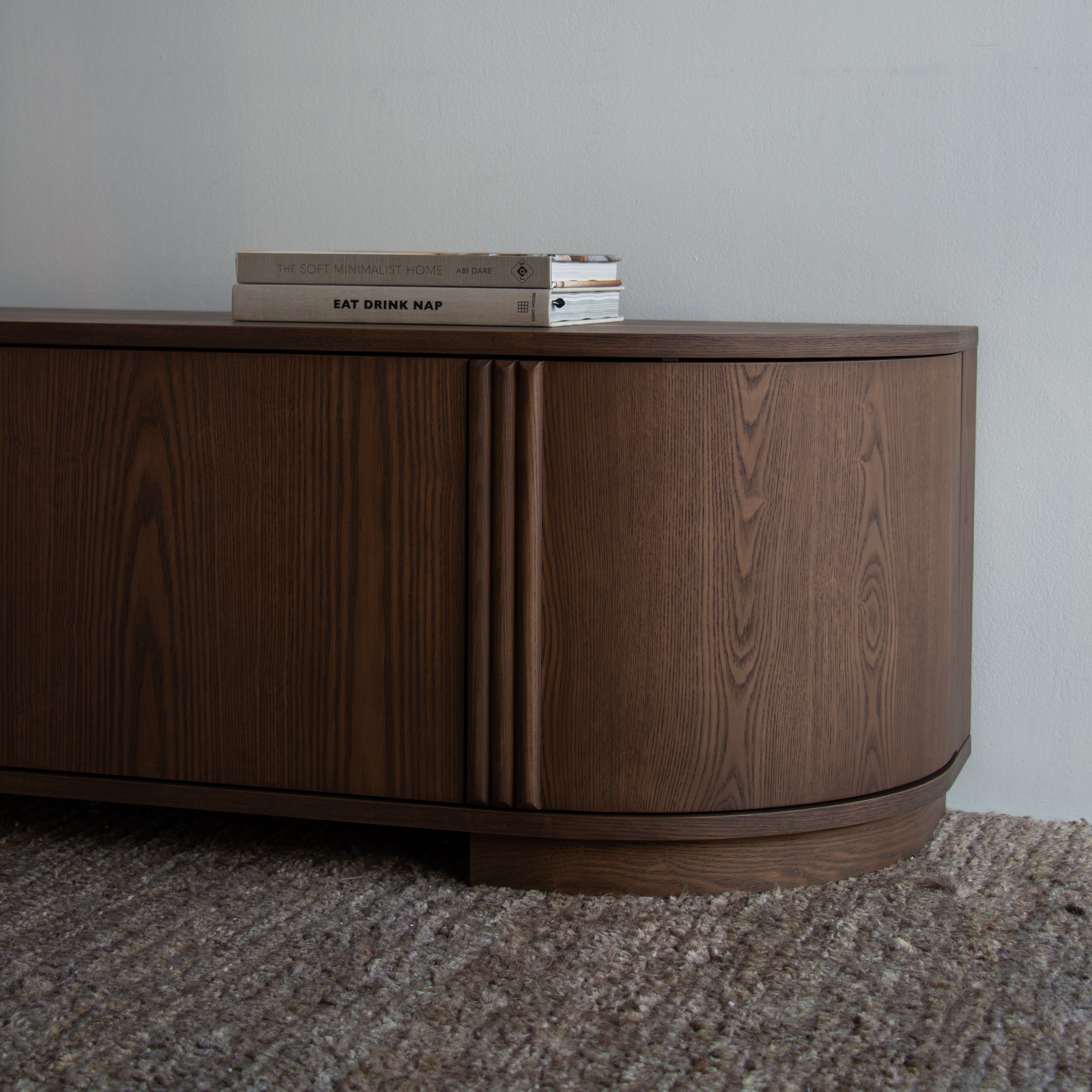 A curved mid-century modern wooden sideboard cabinet with a dark walnut finish, styled on a textured gray carpet against a plain wall with two books on top.