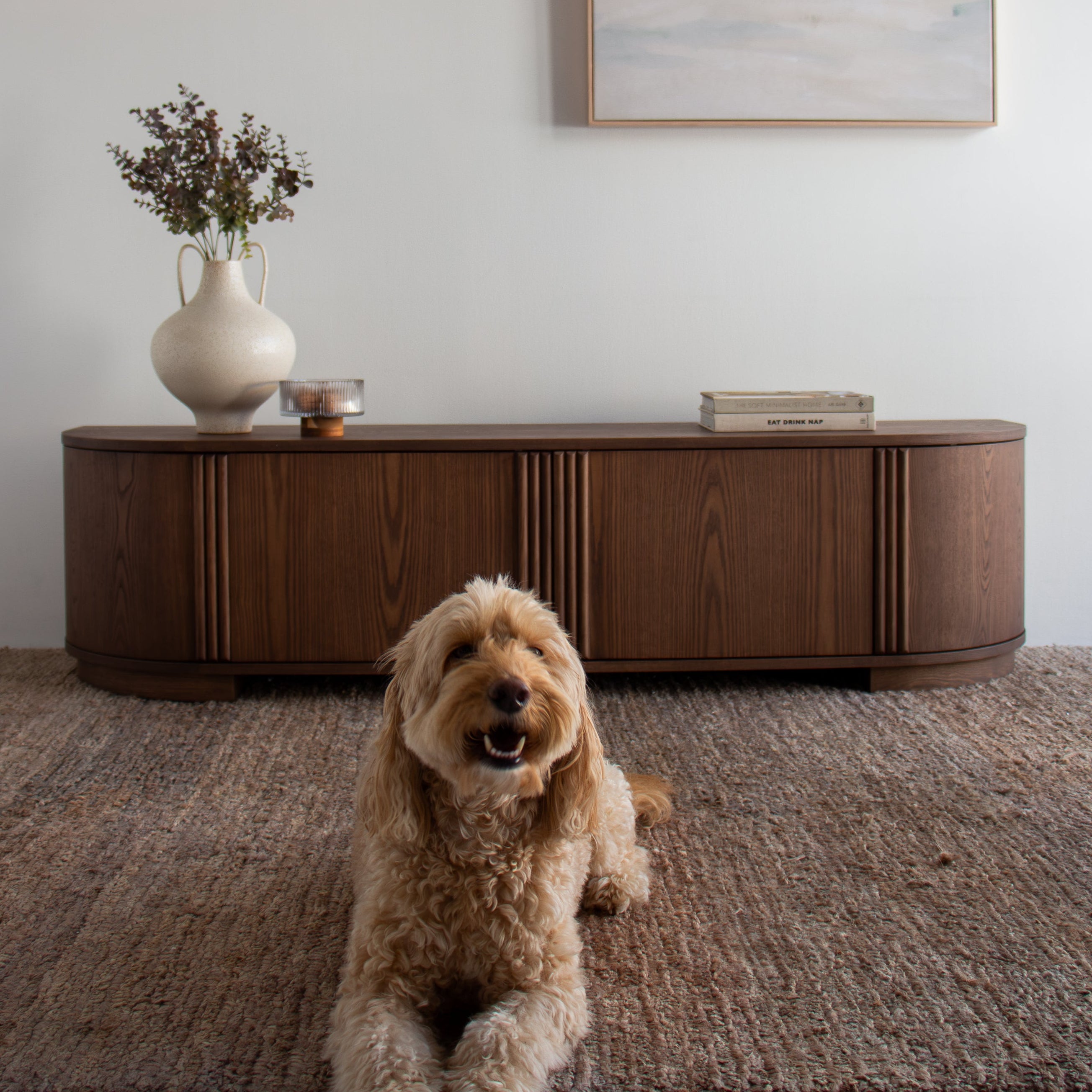 A small fluffy tan dog sits on a textured brown rug in front of a curved dark wood sideboard. On the wall above, a framed abstract landscape painting hangs above the furniture.