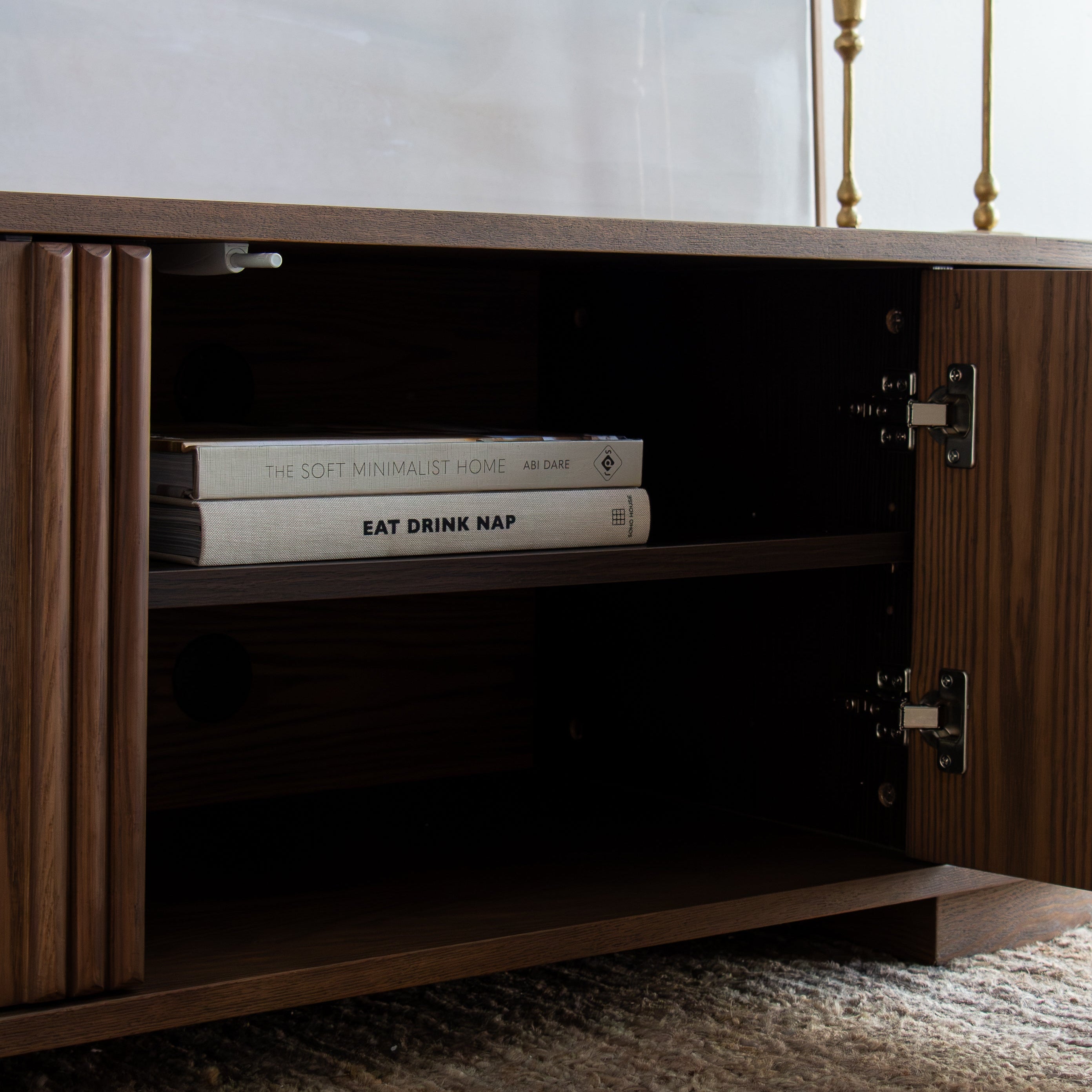 A dark brown wooden media console cabinet with one door open, showing an interior shelf with two books. The cabinet is set on a textured rug, with a blurred modern interior background and two gold candlesticks on top.