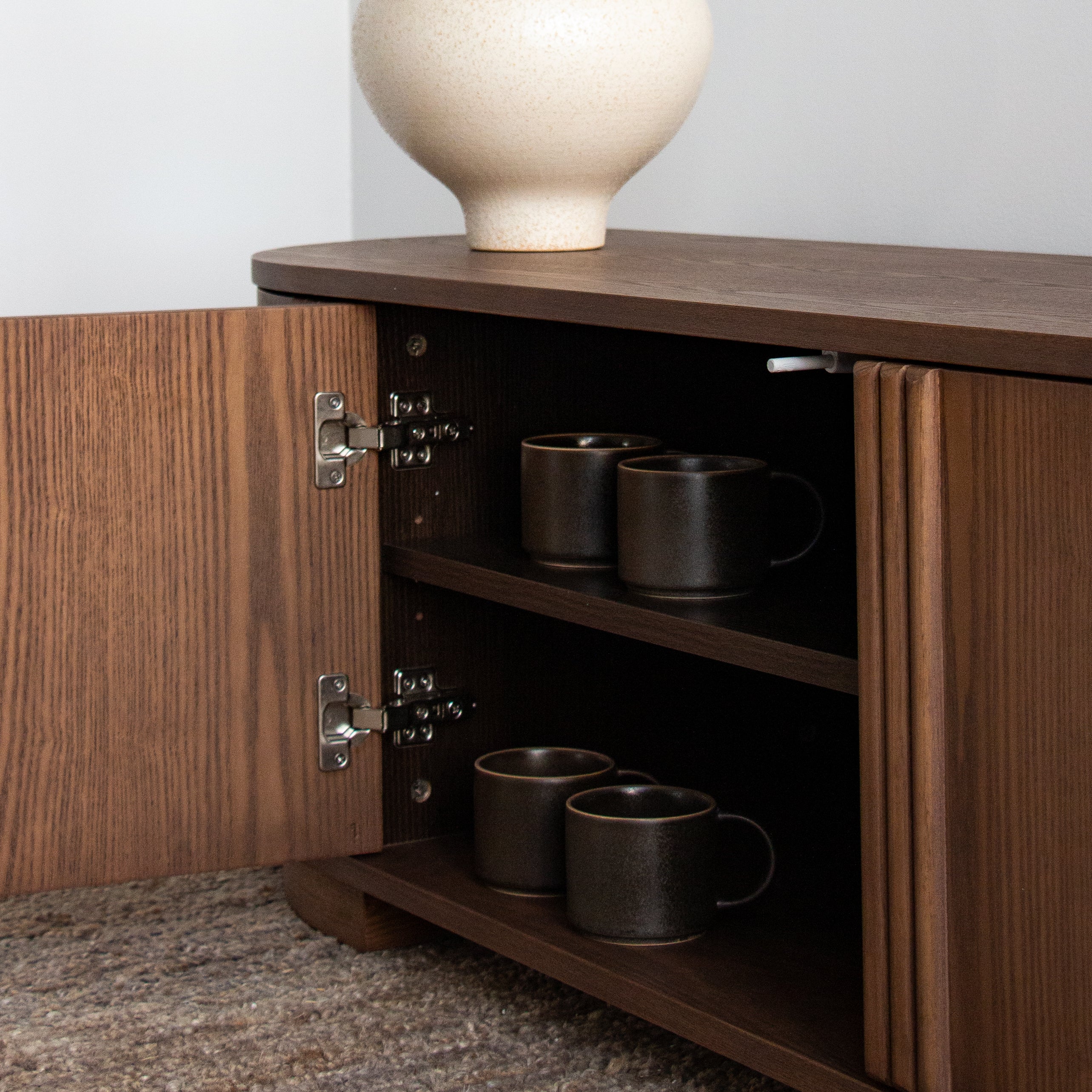 A dark brown wooden sideboard cabinet with one door open, showing two shelves holding four dark ceramic mugs. A large textured cream vase sits on top, and the cabinet is photographed indoors against a light wall with carpet visible at the bottom.