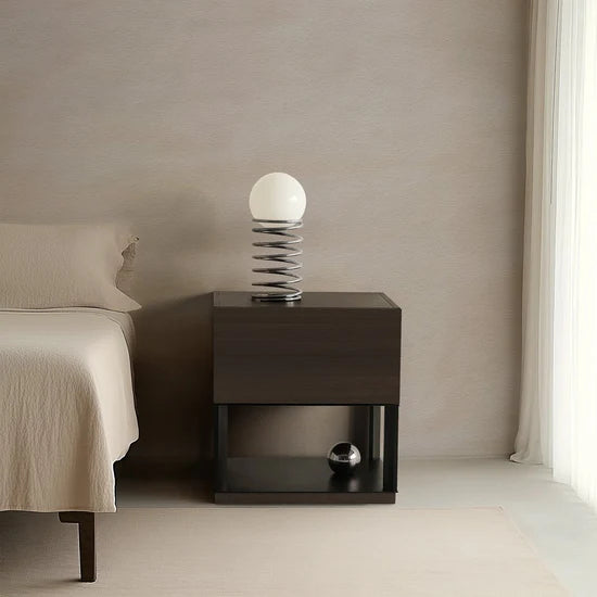 Dark wooden nightstand with decorative lamp next to a bed in a minimalistic bedroom.