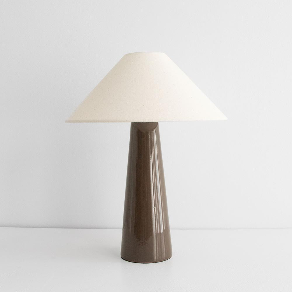 A tapered table lamp with a glossy ceramic base in olive color and a natural linen shade, turned off.