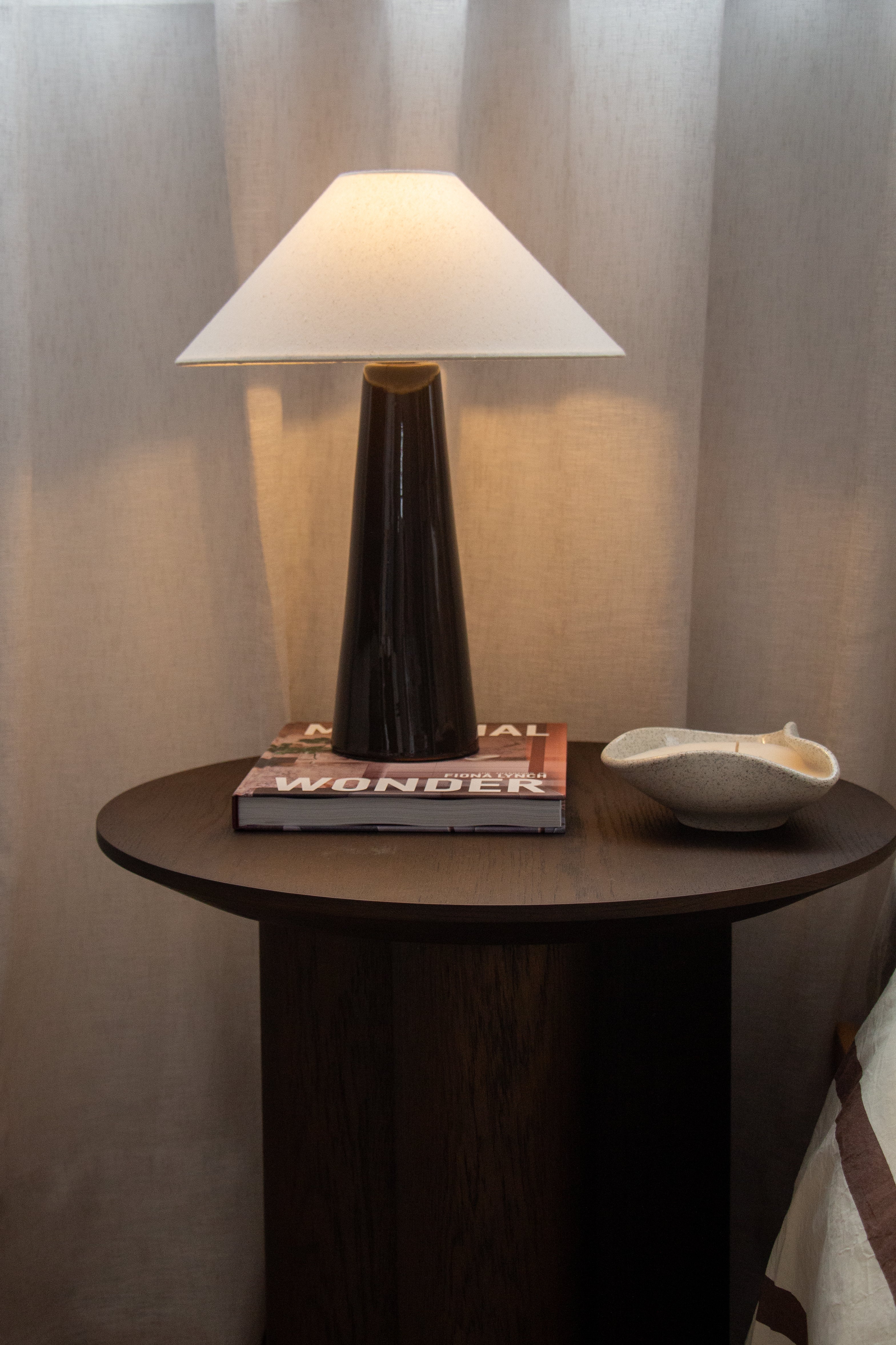 Table lamp on a round wooden table with a book underneath against a neutral wall.