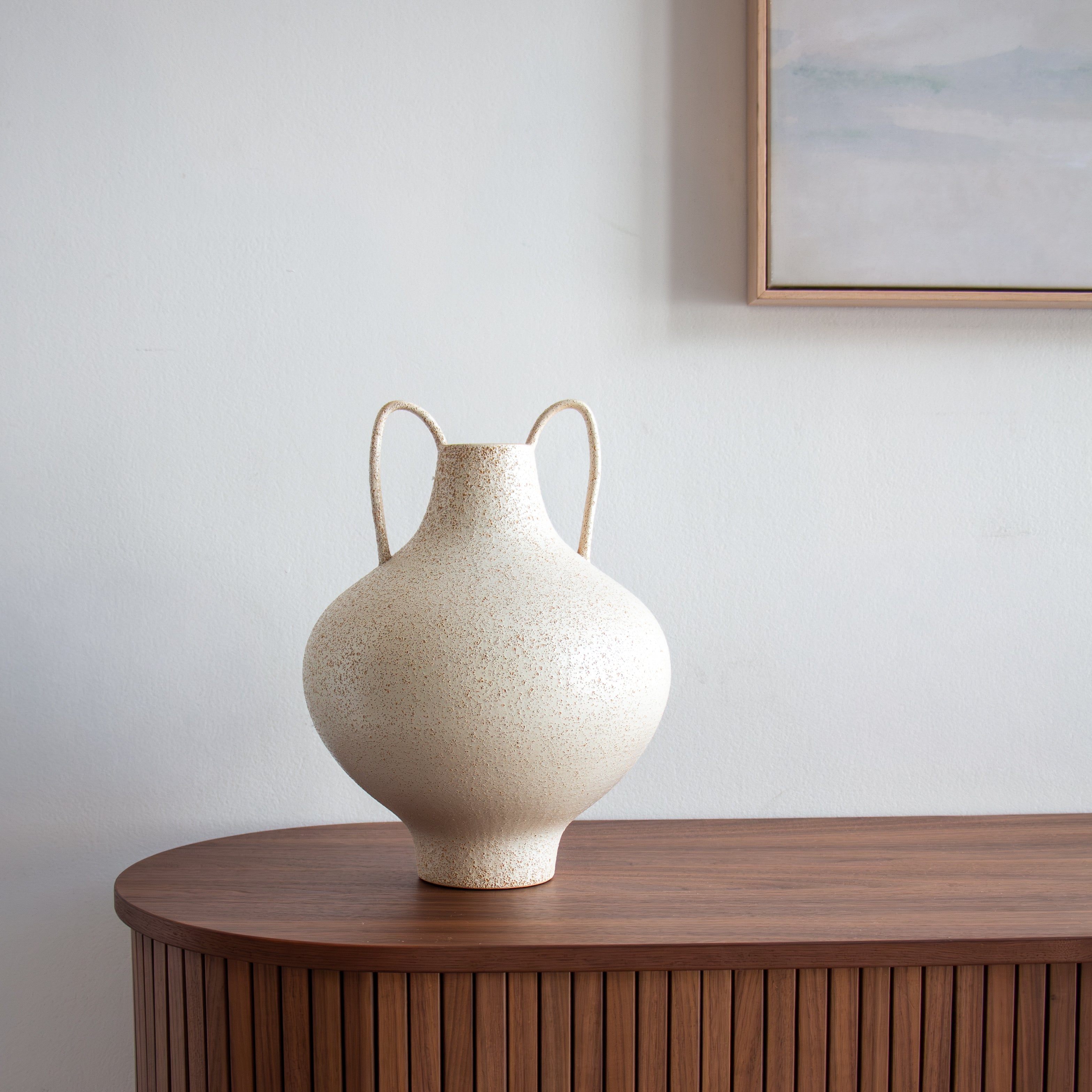 Beige ceramic vase with handles on a wooden surface against a light wall.