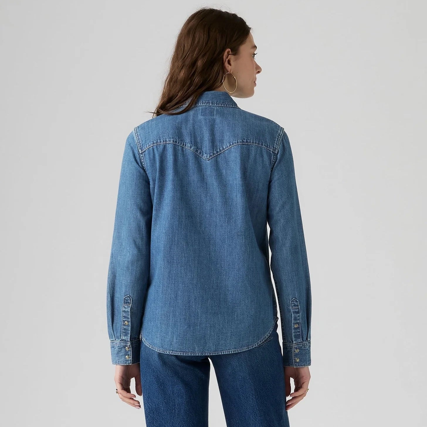 A woman shown from the back wearing a medium-wash blue denim shirt jacket and matching blue jeans against a light gray studio background.