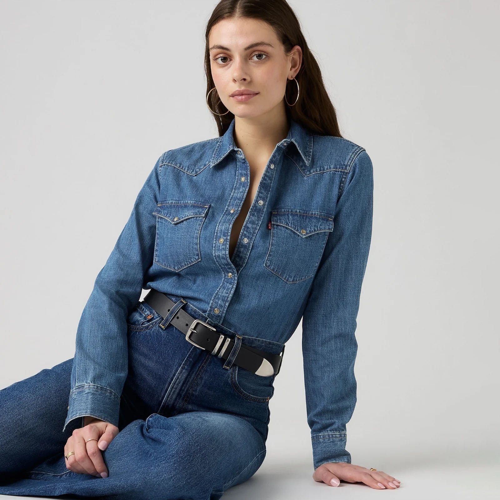 A woman wearing a blue denim long-sleeve shirt with a button-down front, chest pockets, and a matching blue denim skirt, styled with a black belt, seated against a light gray studio background.