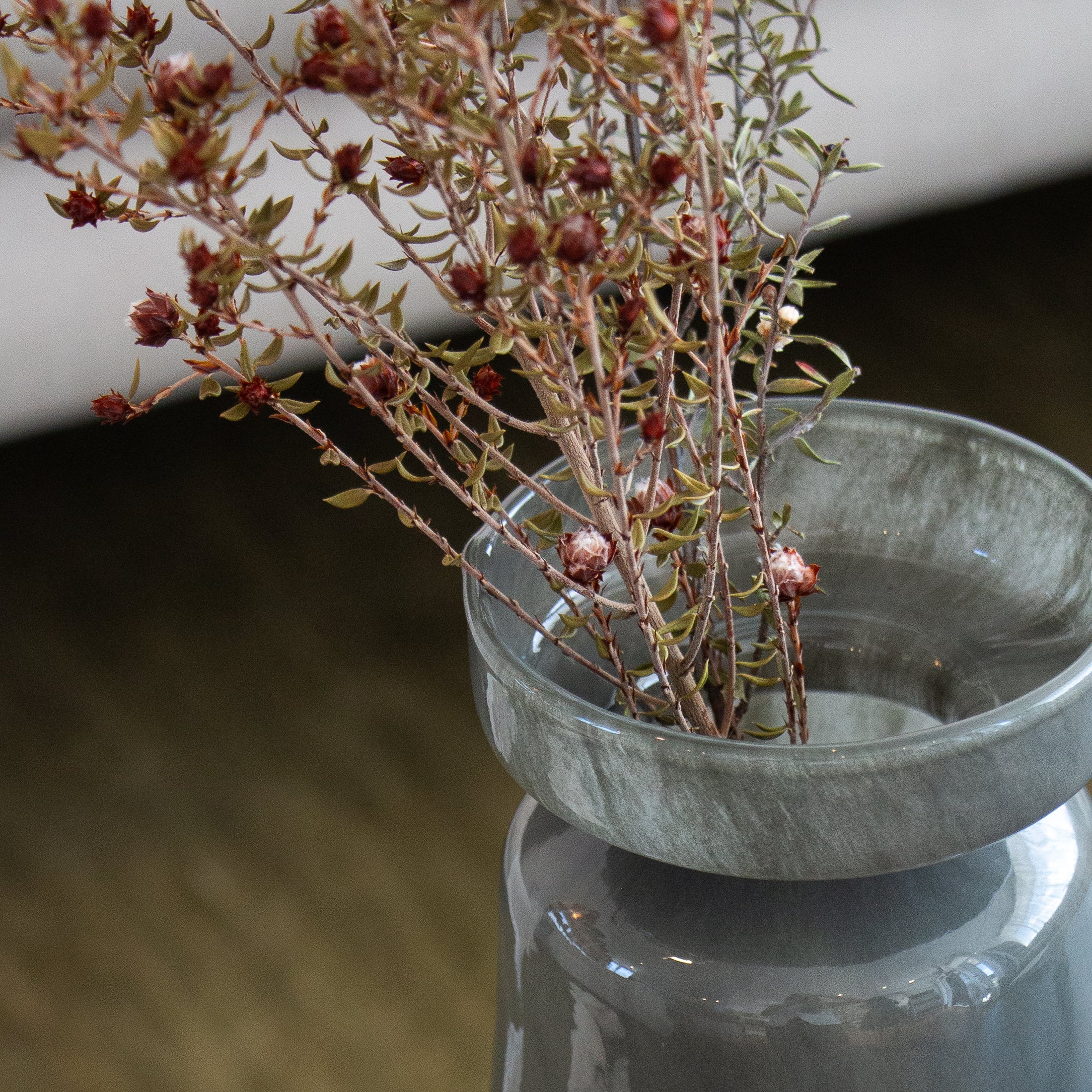 Dried plant in a dark green glass vase on a reflective surface with a blurred background