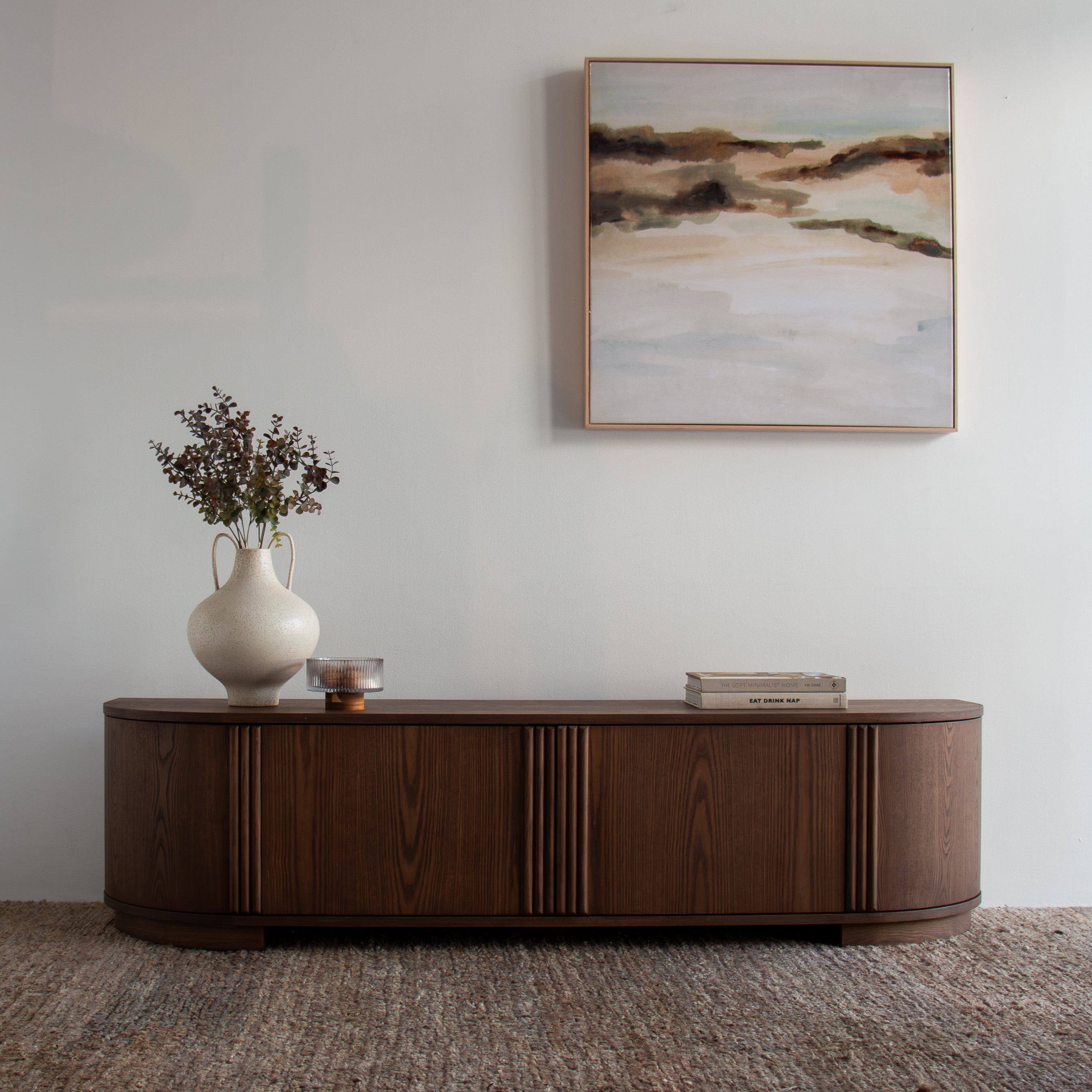A curved dark wood sideboard or credenza in a styled room setting, featuring a white ceramic vase with dried flowers, a small metallic bowl, and a stack of books on top. Above the sideboard, a framed abstract landscape painting hangs on a light gray wall, and a textured brown rug covers the floor.