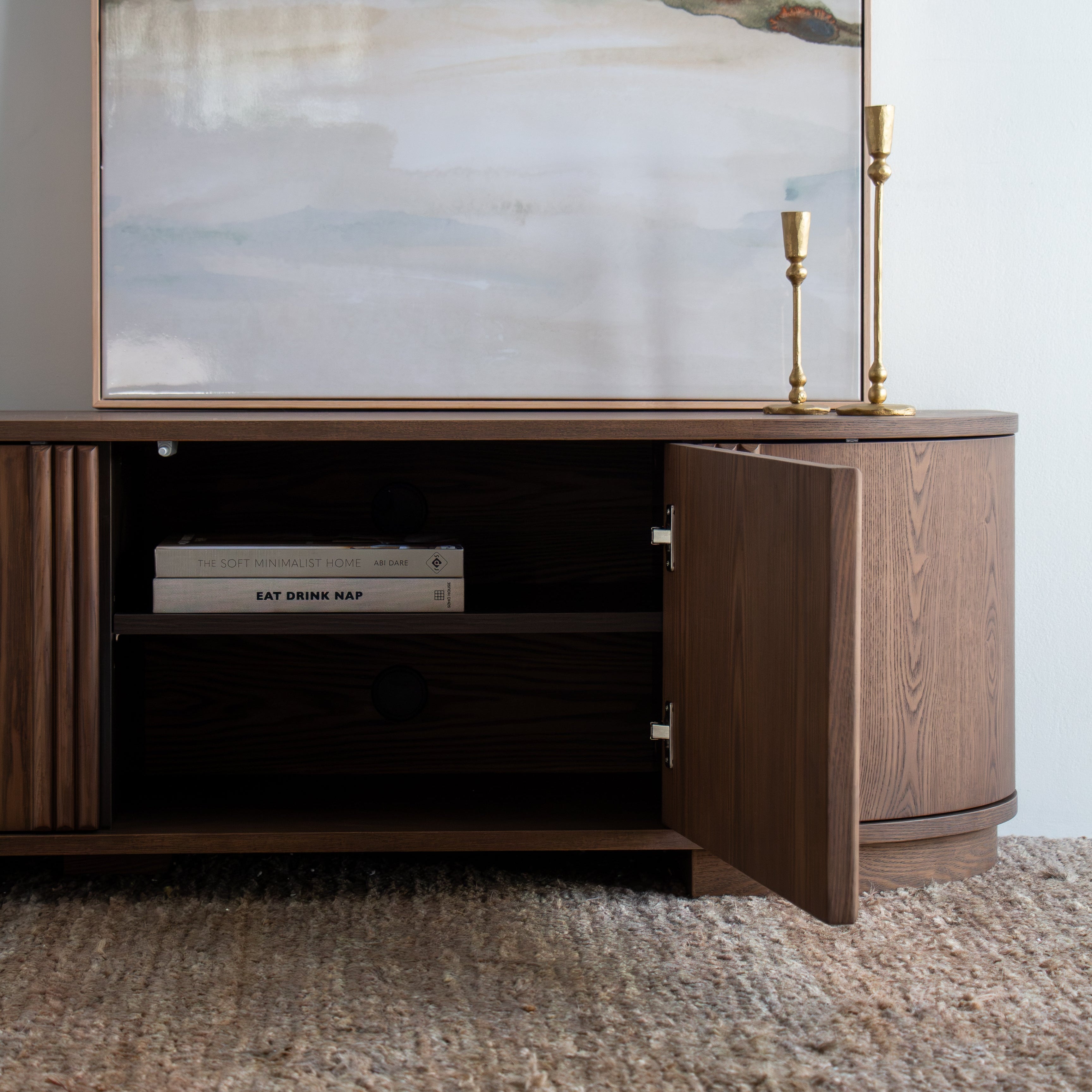 A modern dark wood media console with one door open, styled in a room setting with a large abstract landscape painting above and a textured rug underneath.