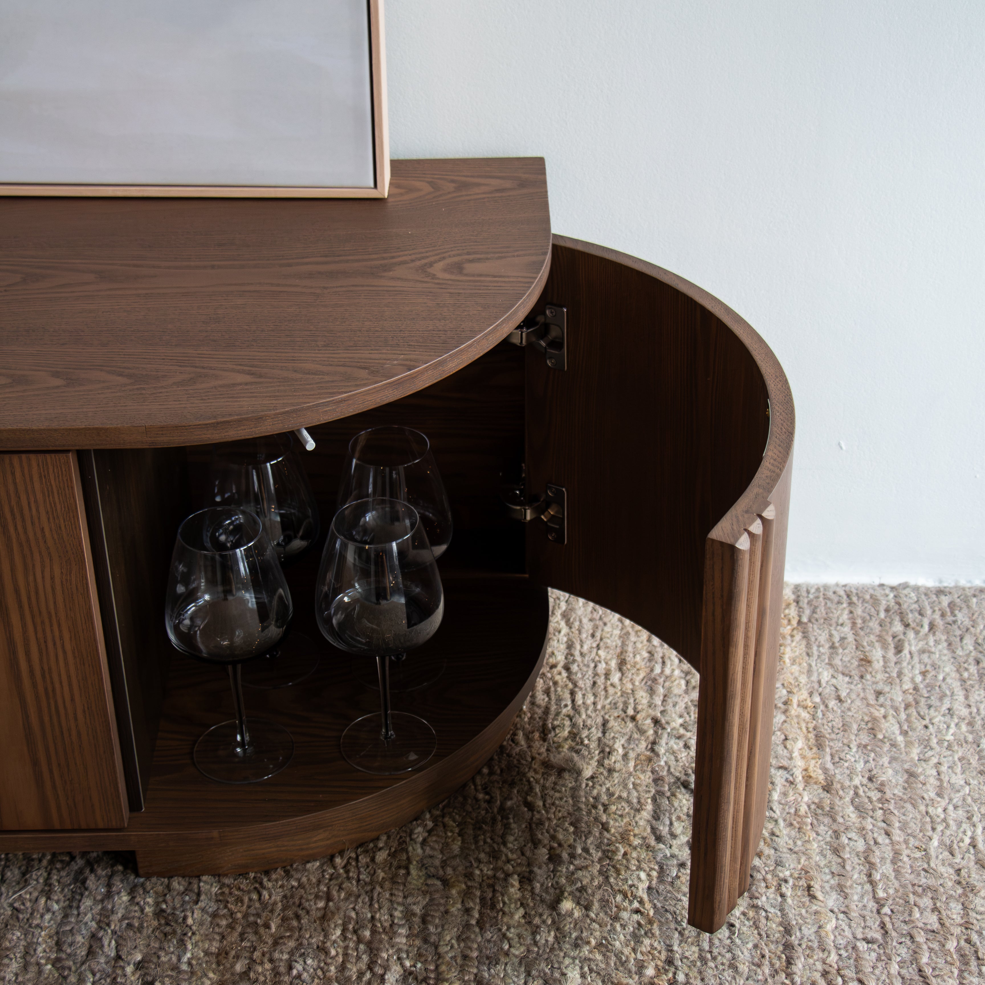 A curved dark wood cabinet with one door swung open, revealing wine glasses inside, set on a textured beige rug against a white wall.