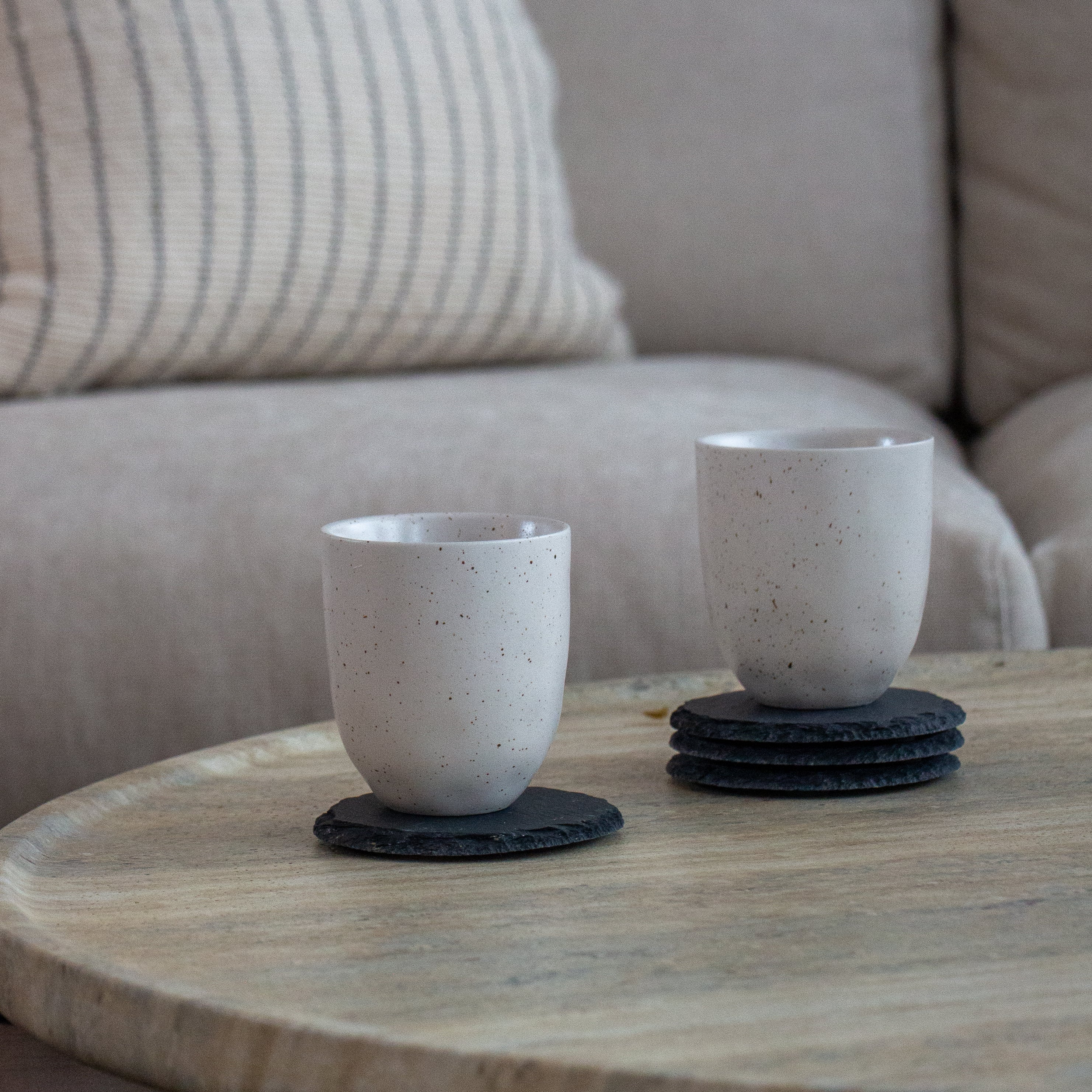 Two ceramic cups on coasters on a wooden table with a couch in the background