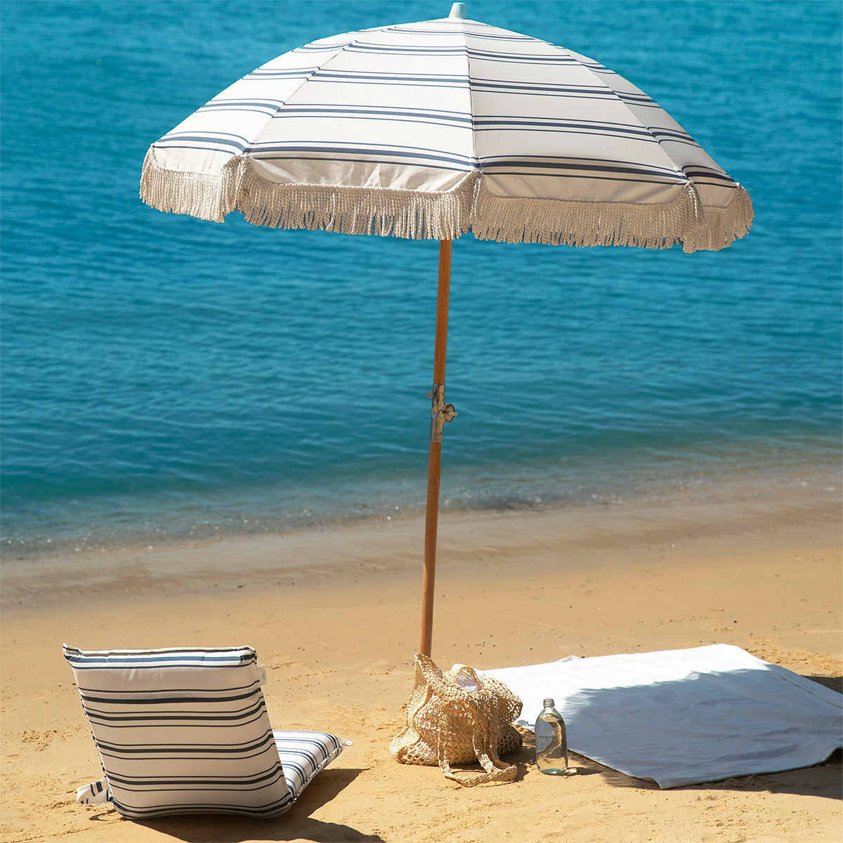 A beach umbrella with vintage stripes in a coastal blue and white color scheme, with a wooden pole and aluminium tilt function, set up on a beach with a carry bag beside it.