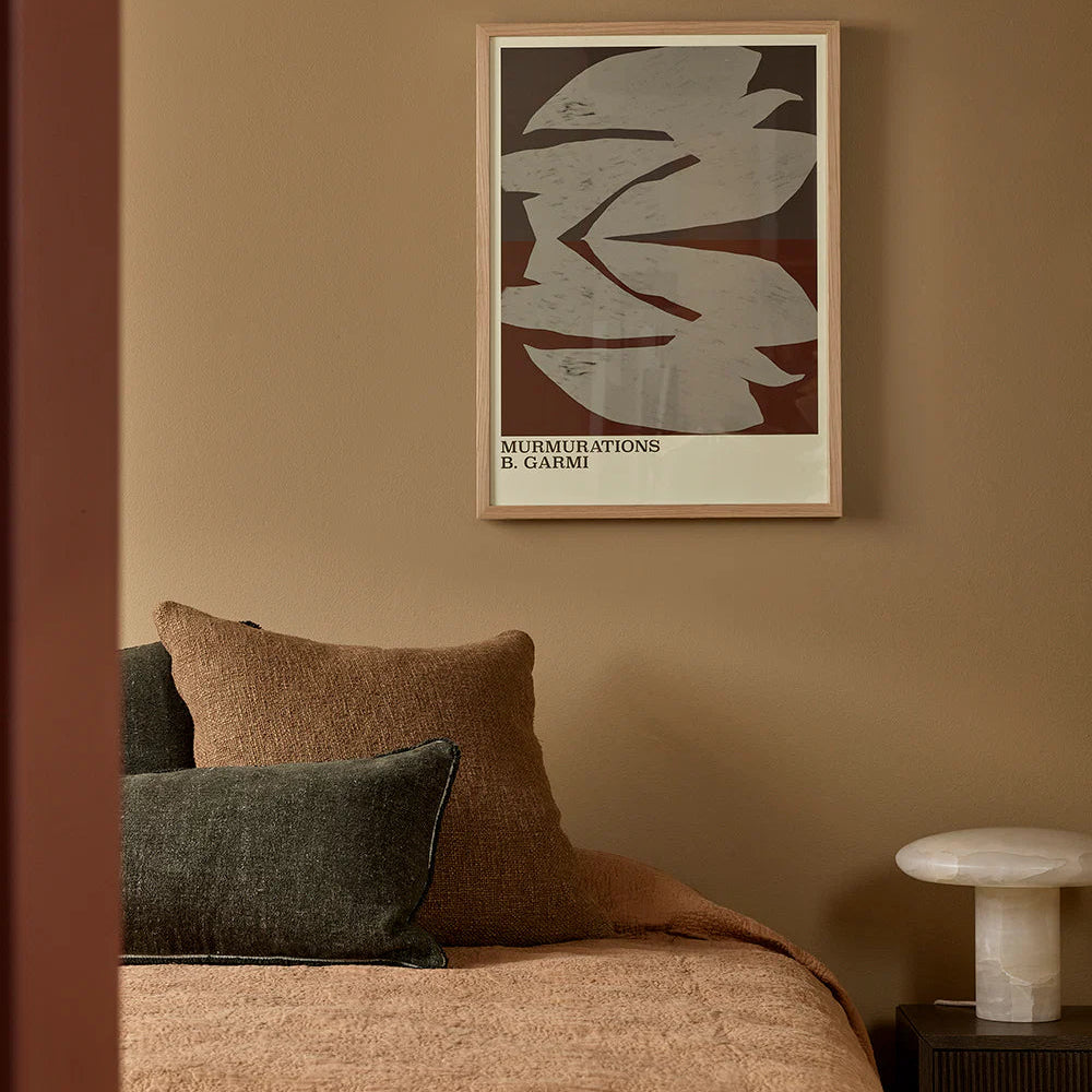 Framed artwork on a wall above a bed with pillows and a lamp.